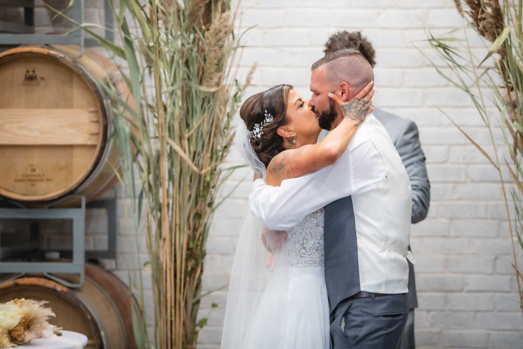 A bride and groom kiss during their wedding ceremony in front of barrels.