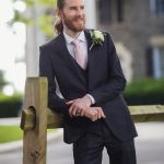 A man in a wedding suit leaning against a fence.
