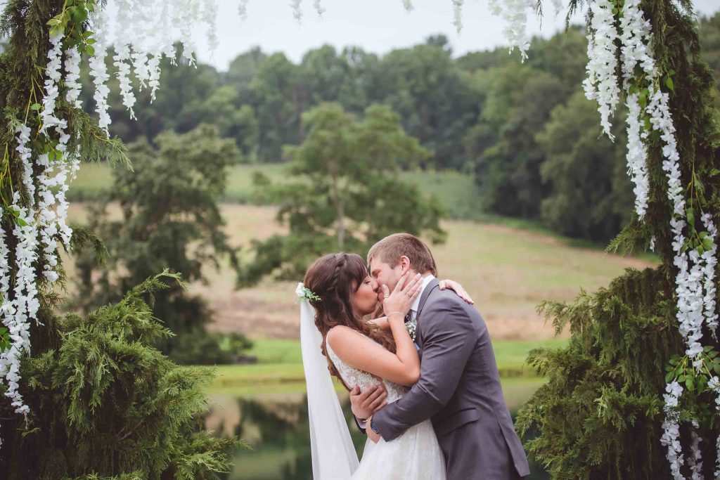 A bride and groom share a romantic kiss during their wedding ceremony by a pond.