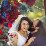 A portrait of a bride and groom kissing under a red and blue wedding arch.