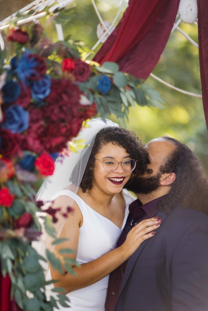A portrait of a bride and groom kissing under a red and blue wedding arch.
