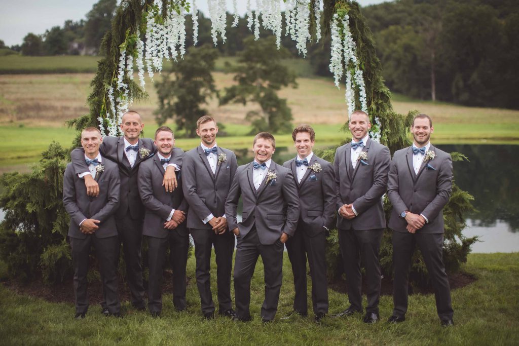 Groomsmen in gray tuxedos standing in front of a wedding arch.