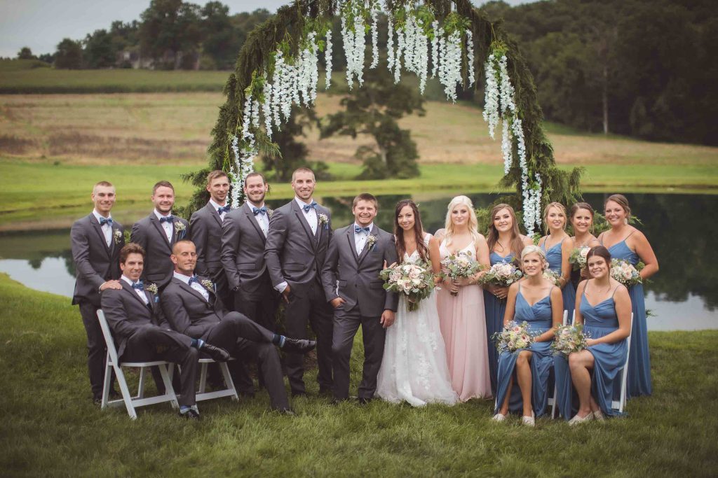 A group of bridesmaids and groomsmen pose for a wedding portrait in front of an arch.