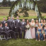 A group of bridesmaids and groomsmen pose for a wedding portrait in front of an arch.