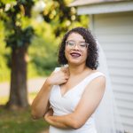 A smiling bride in a white wedding dress posing in front of a house for her portrait.