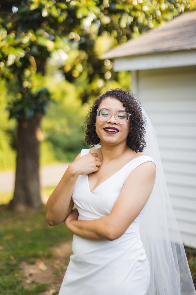 A smiling bride in a white wedding dress posing in front of a house for her portrait.