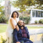 A wedding portrait of a bride and groom posing on a bench in front of a tree.