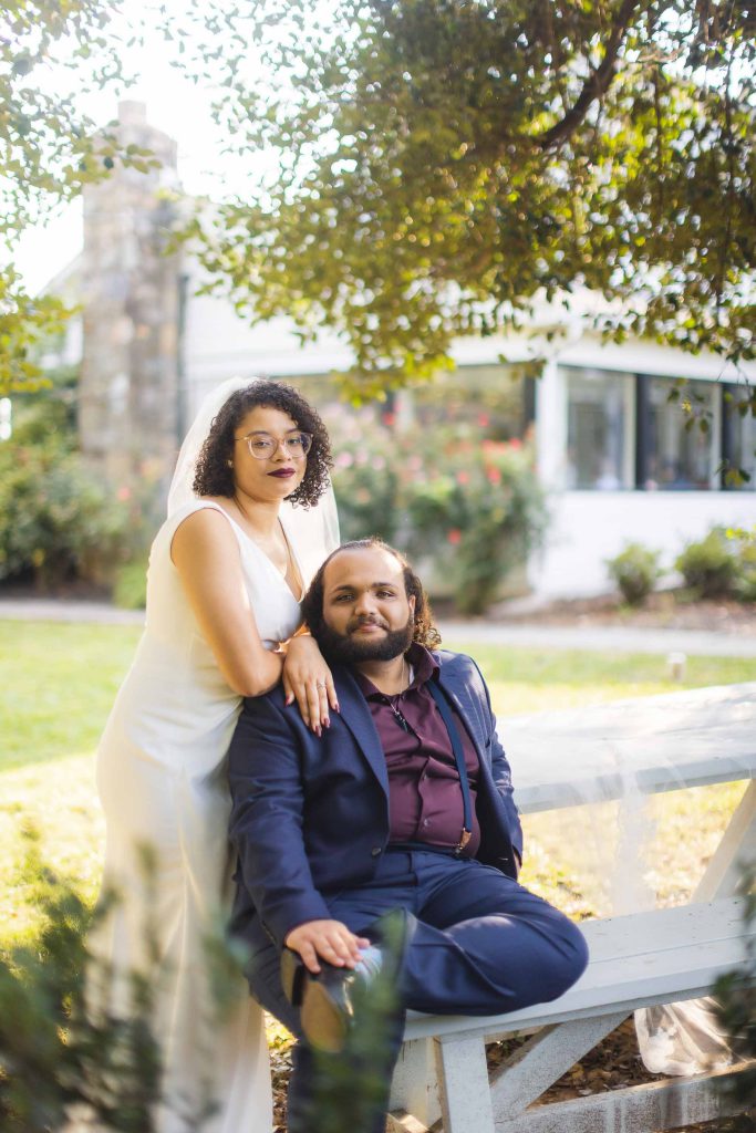 A wedding portrait of a bride and groom posing on a bench in front of a tree.