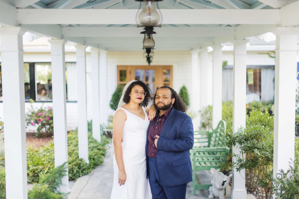 A wedding portrait of a bride and groom standing under a covered porch.