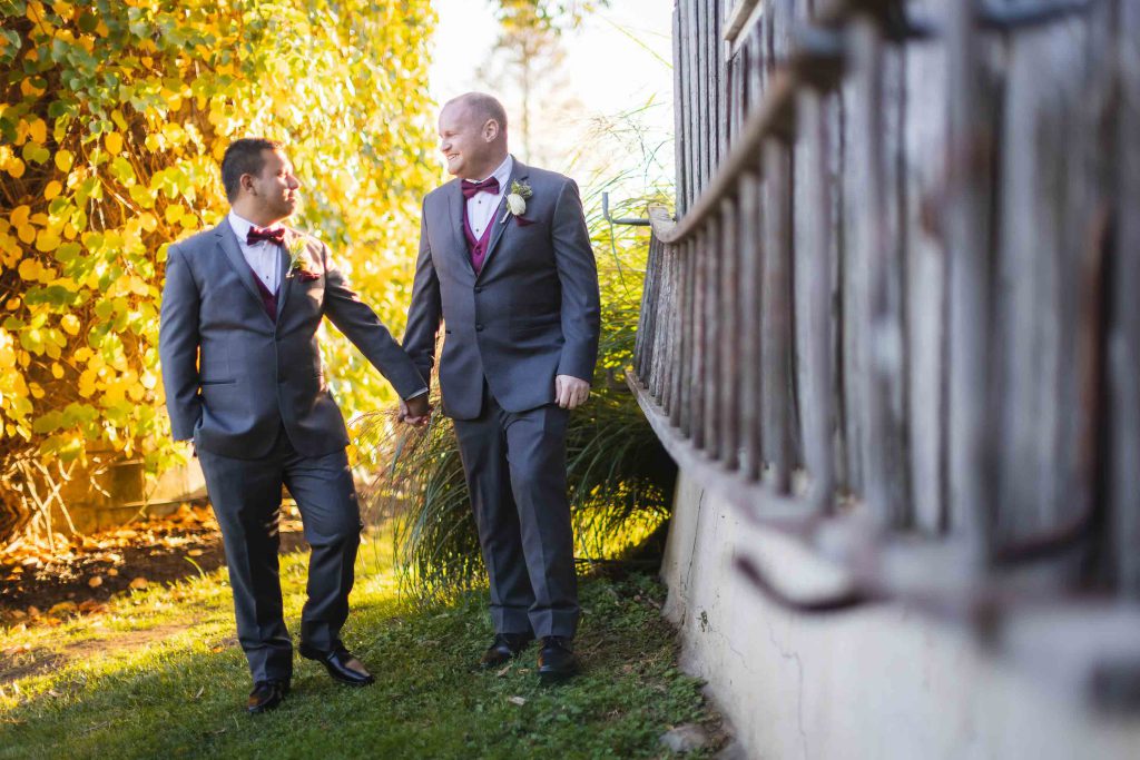 Two grooms holding hands in front of a fence, capturing their wedding portrait.