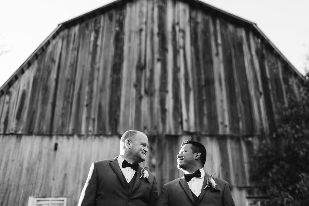 Wedding portrait of two grooms standing in front of a barn.
