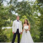 A wedding portrait of a bride and groom in a white tuxedo standing on a stone wall.