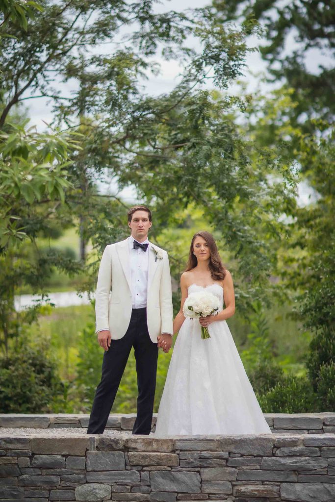 A wedding portrait of a bride and groom in a white tuxedo standing on a stone wall.