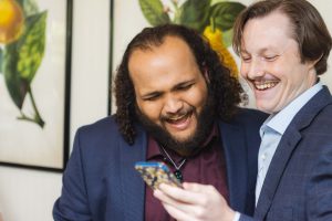 Two men are smiling at a wedding while holding a cell phone.