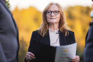A woman wearing glasses is holding a book in front of a group of people during a wedding ceremony.