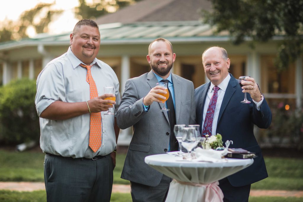 Three men in suits standing next to a table at a wedding reception with drinks.