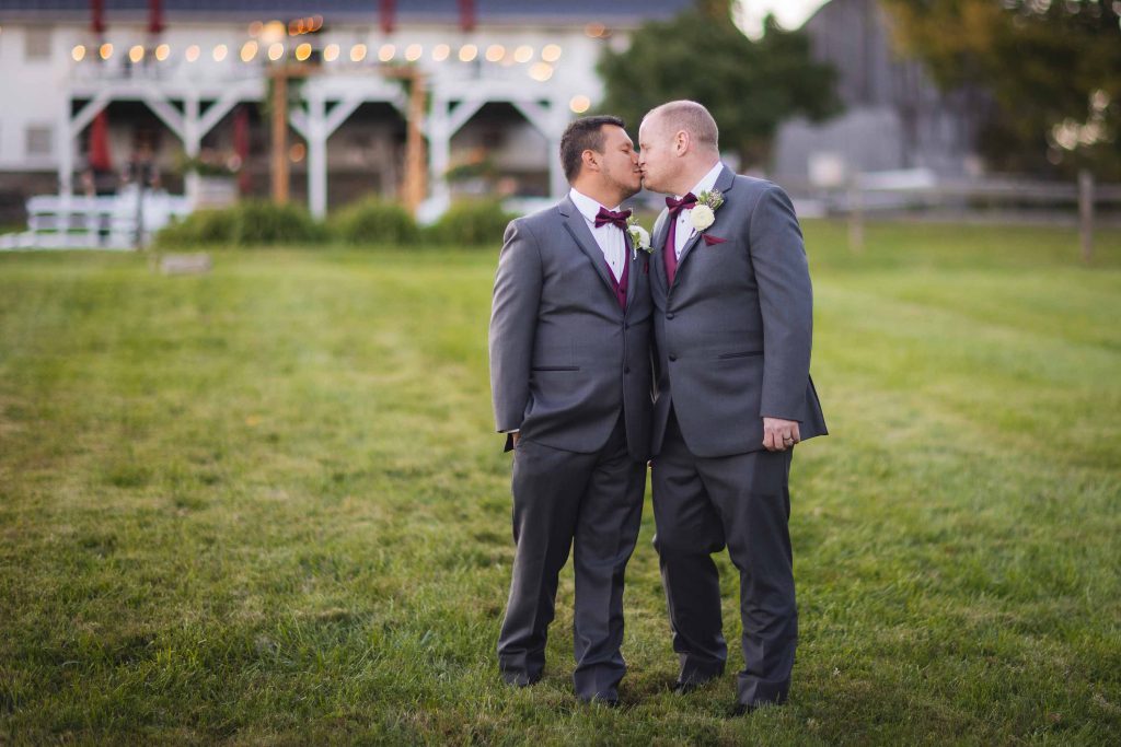 Wedding portrait of two grooms kissing in front of a barn.