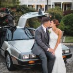 A couple in a wedding portrait kiss in front of a Delorean car.