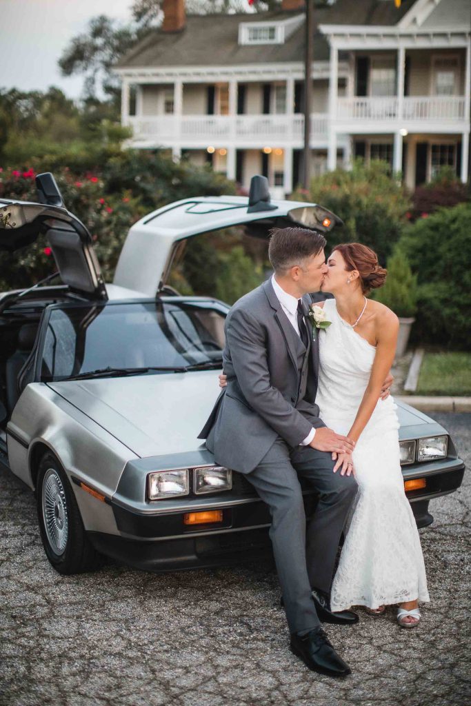 A couple in a wedding portrait kiss in front of a Delorean car.