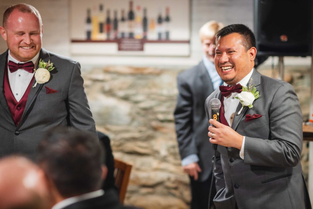 Two men in suits laughing at each other at a wedding reception.