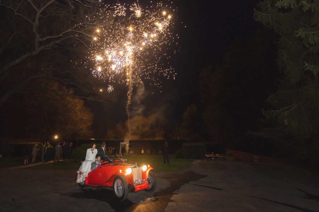 A couple celebrating their wedding with a grand exit in a red car accompanied by fireworks.
