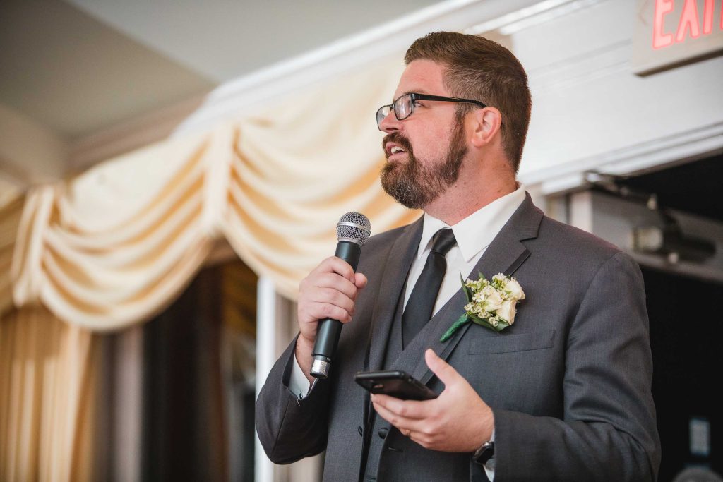 A man in a suit speaking at a wedding reception.