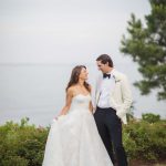 A wedding portrait of a bride and groom standing in front of the ocean in a tuxedo.