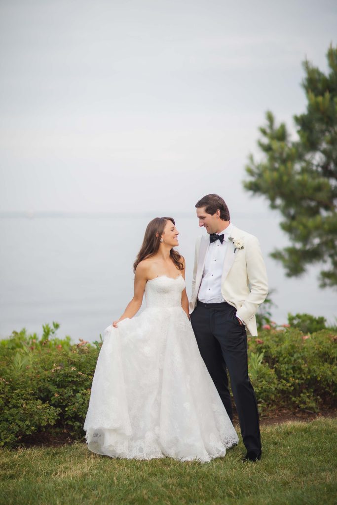 A wedding portrait of a bride and groom standing in front of the ocean in a tuxedo.