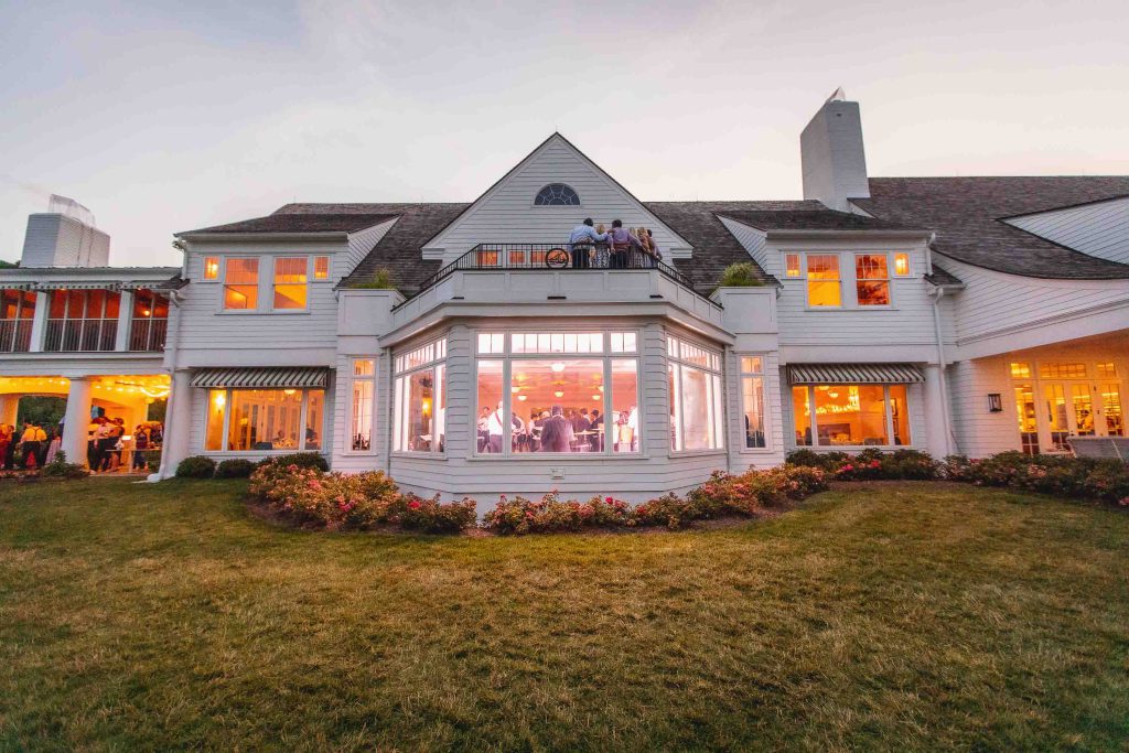 A white house with people sitting on the porch, perfect for a wedding reception at dusk.