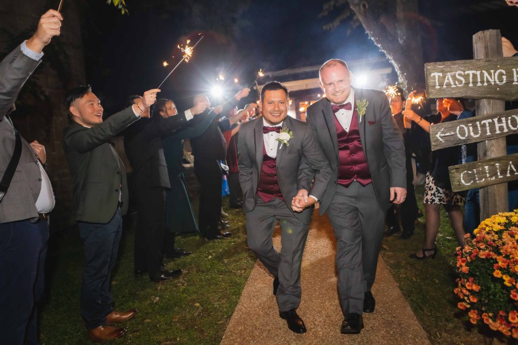 Two wedding grooms walking down a path with sparklers in their hands during the reception.