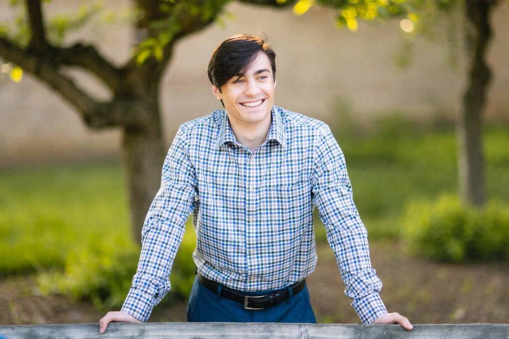 A young high school graduate leaning against a fence in a park.