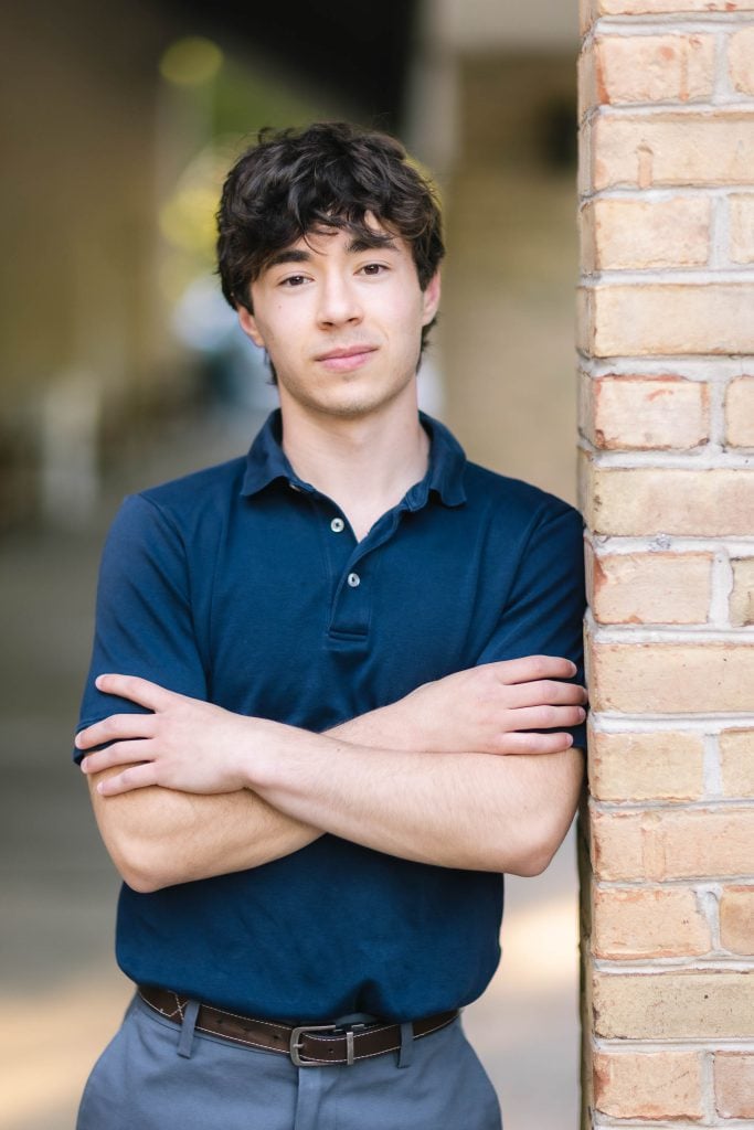 A senior high school student in a blue shirt leaning against a brick wall.