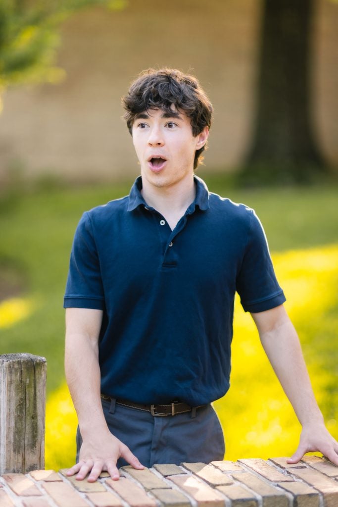 A young man in a blue polo shirt leaning against a brick wall for his graduation portrait.