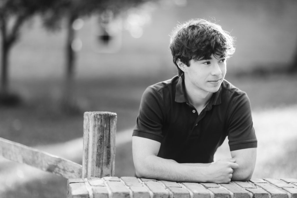 A black and white portrait of a senior high school student leaning against a brick wall.