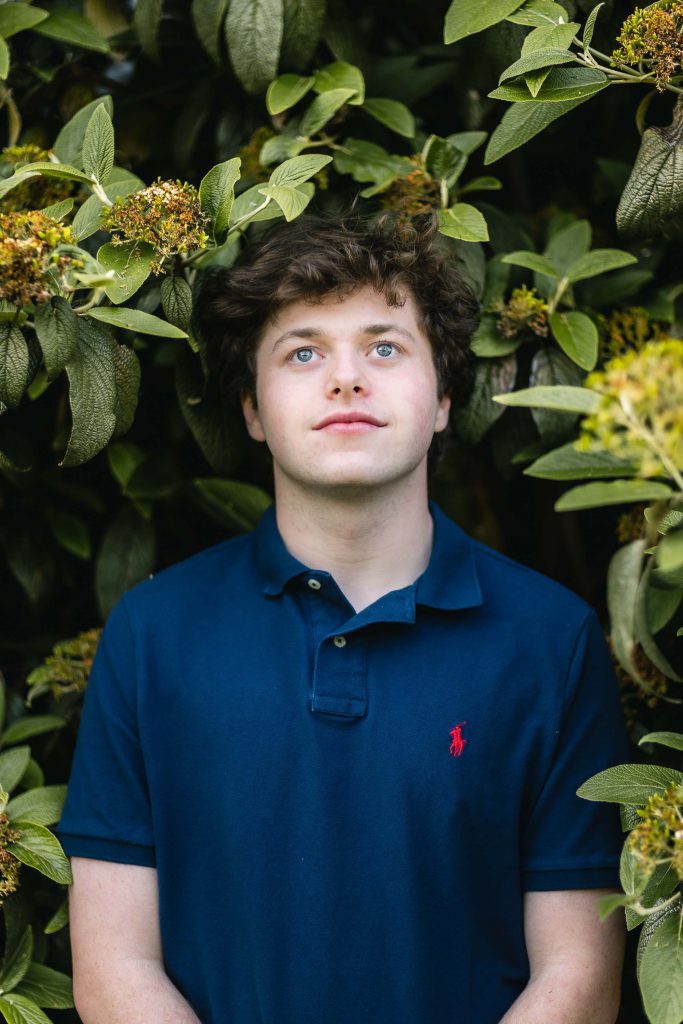 A young senior in a blue polo shirt standing in front of bushes.