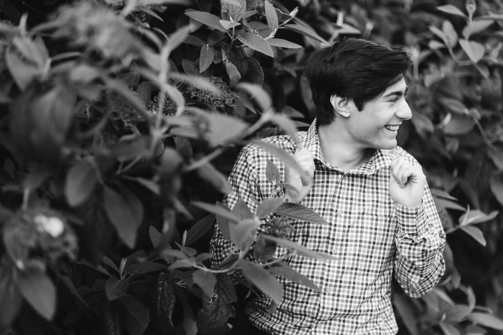 A black and white photo of a high school senior leaning against a bush.