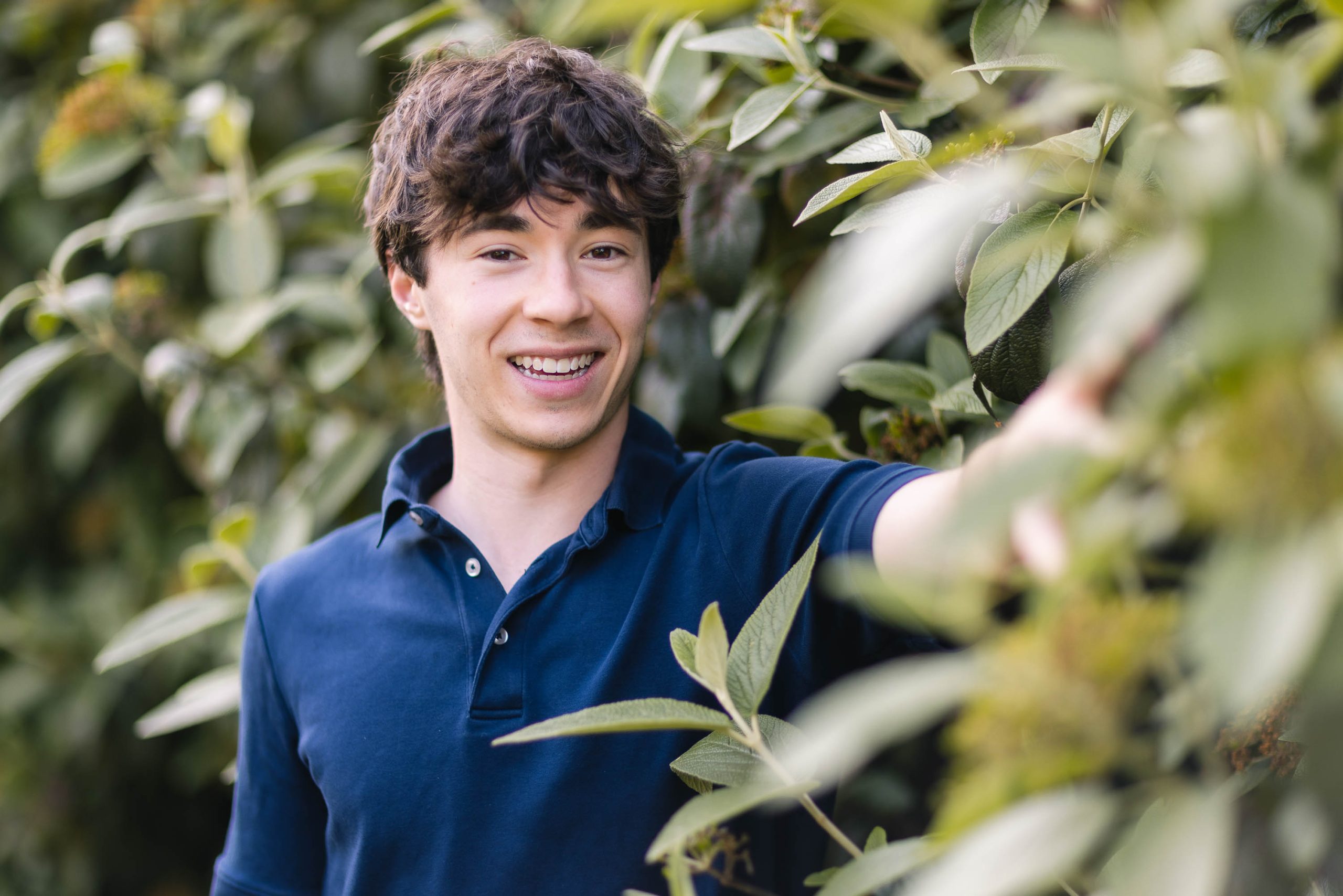 A young man in a blue shirt standing next to a bush for his senior portrait.