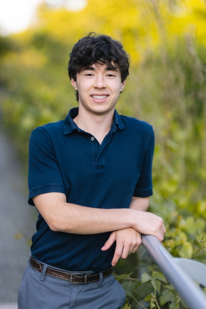 A senior young man in a blue shirt leaning against a railing.