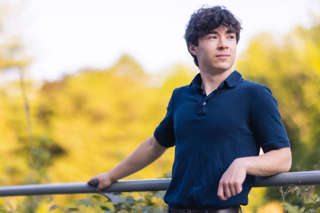 A young man in a blue shirt at his high school graduation leaning against a railing.