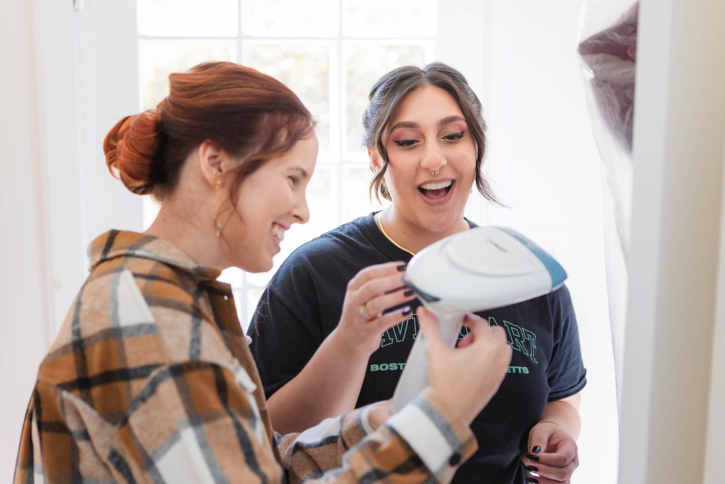 Two women candidly laughing while preparing for a wedding, using a hairdryer.