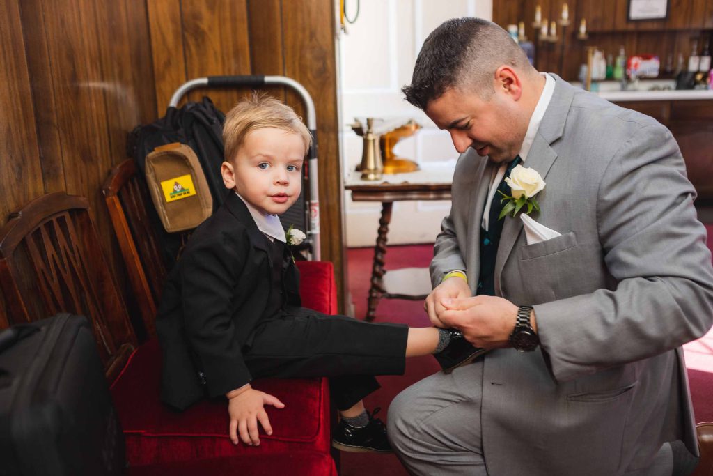 A candid man in a suit helping with wedding preparation by putting a boy's shoe on.