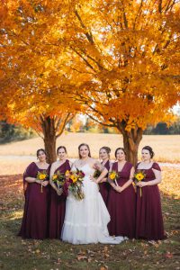 Bridesmaids in burgundy posing for a wedding portrait in front of a fall tree.