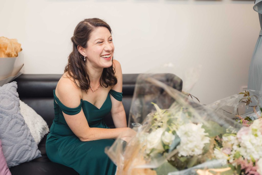 A woman in a green dress candidly laughs while sitting on a couch during wedding preparation.