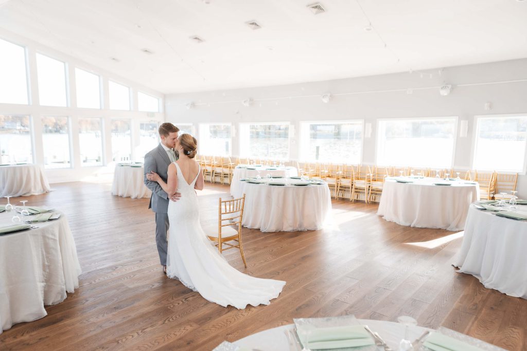 A candid bride and groom dance during wedding preparations in a room with large windows.