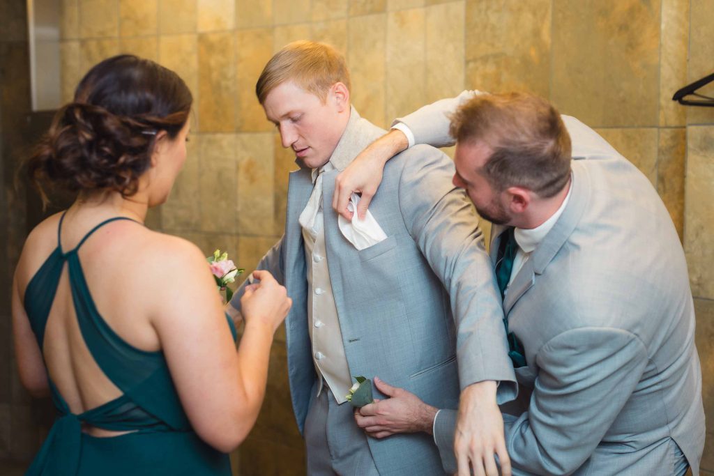 A man in a suit is candidly preparing another man for a wedding by helping him put on a boutonniere.