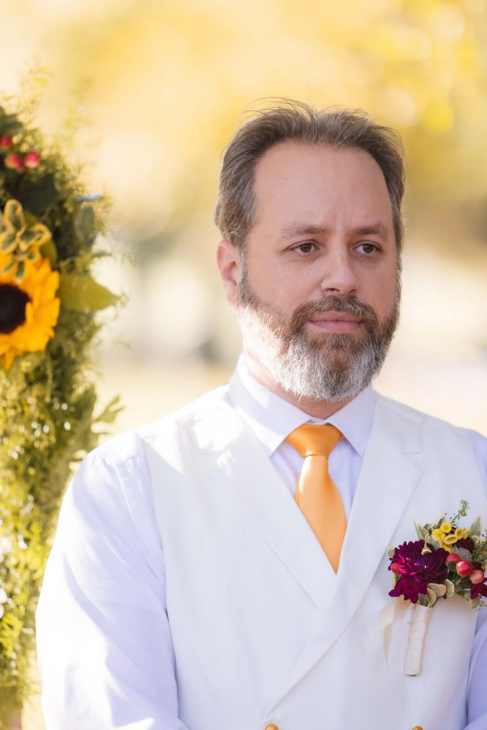 A man in a white vest and yellow tie is standing in front of sunflowers at a ceremony.