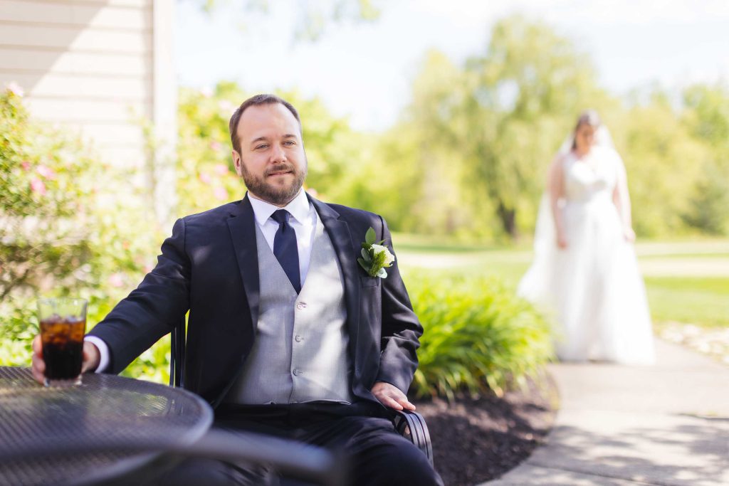A man in a suit sits at a table with a drink during a wedding.