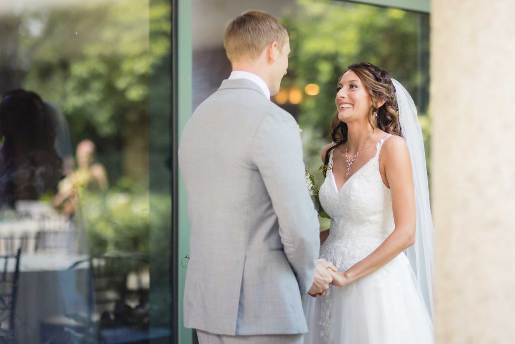 A candid wedding couple looks at each other in front of a glass door as part of their preparation.
