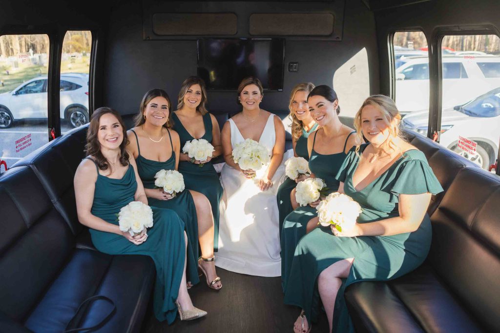 A group of bridesmaids in candid wedding preparation sitting on the back of a limo.