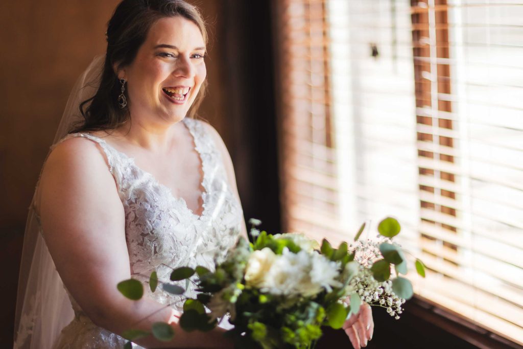 A bride smiles holding her bouquet in front of a window for a wedding portrait.